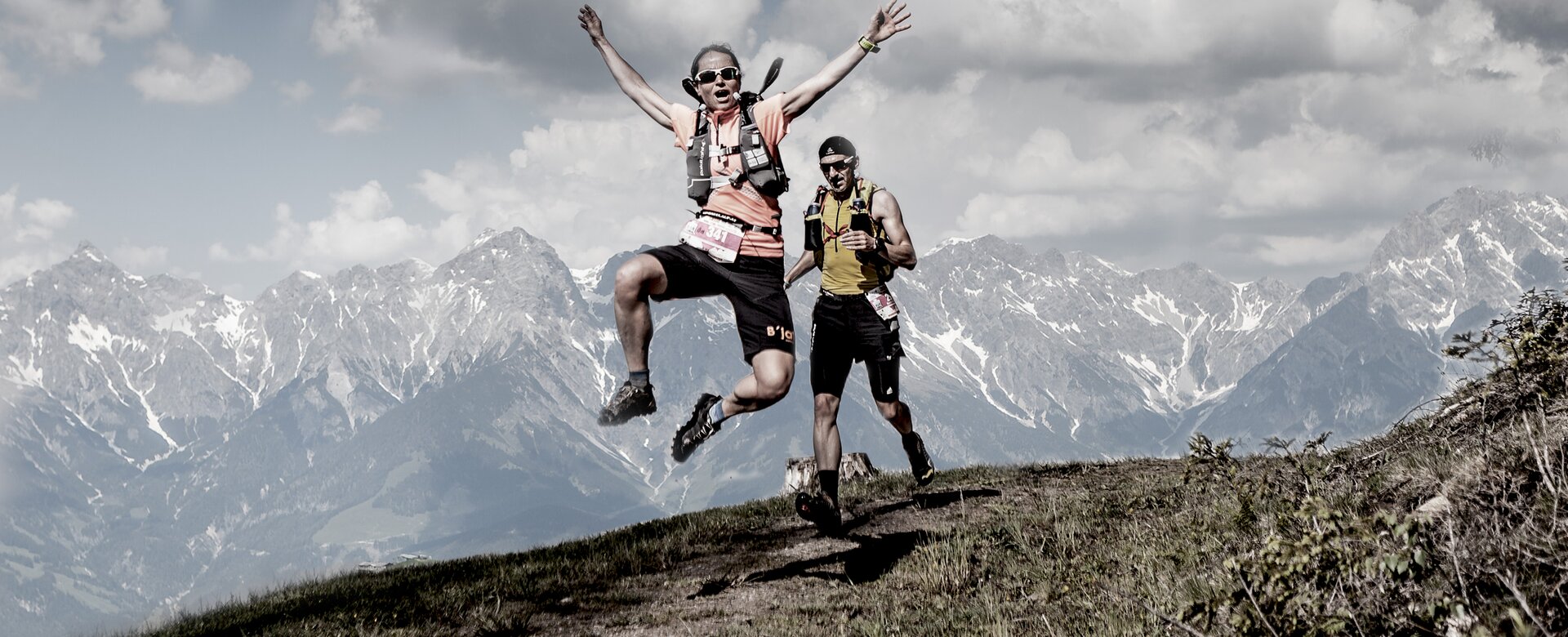 Zwei Trailrunner laufen am Bergkamm, einer springt mit ausgestreckten Armen vor der Kulisse schneebedeckter Berge. | © Miriam Lottes