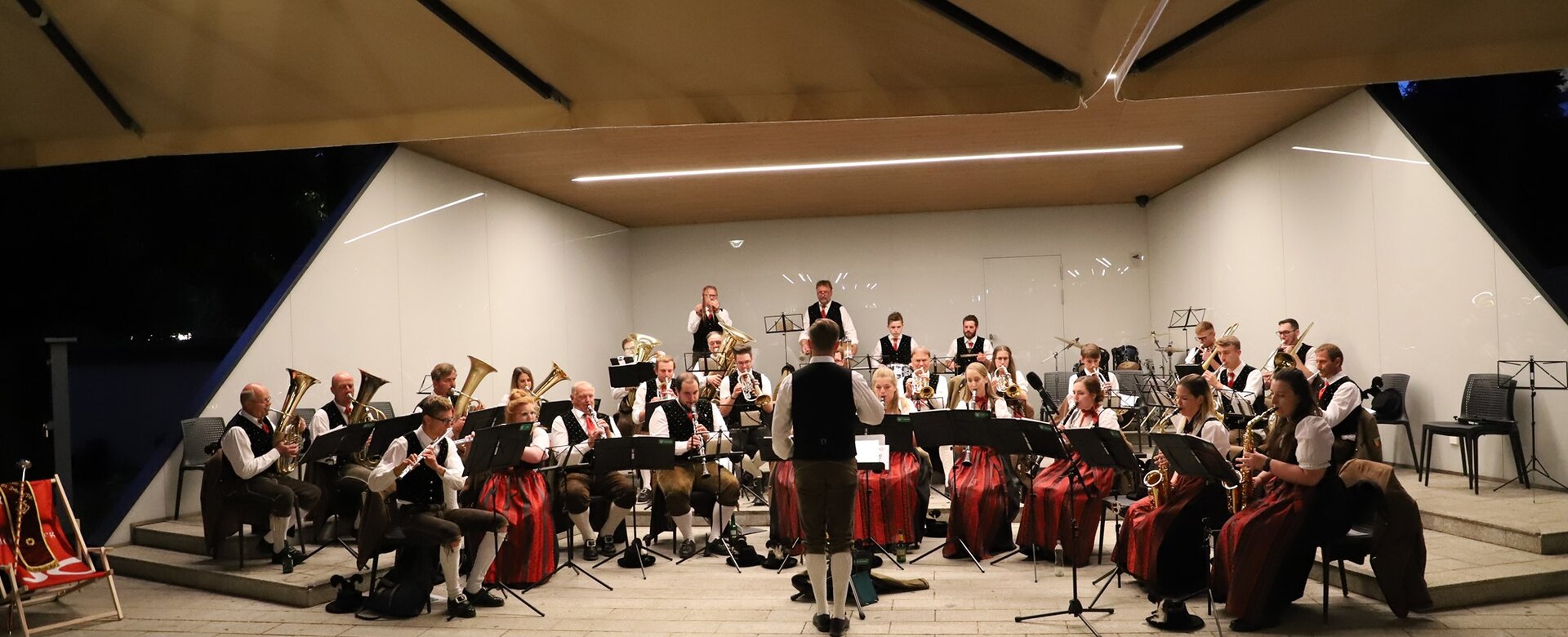 Blasorchester in Tracht bei abendlichem Stadtkonzert auf überdachter Freiluftbühne, Dirigent steht vorn | © Bürgermusik