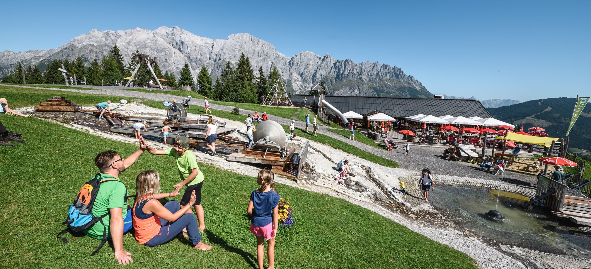 Familien genießen den Wasserspielplatz an der Karbachalm vor imposanter Hochkönig-Kulisse bei Sonnenschein