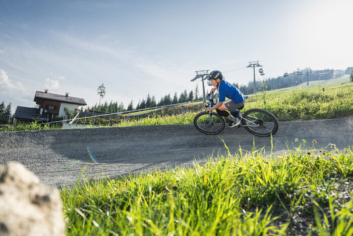 Kid rides Woom bike past alpine house and summer gondola lift. | © Snow Space Salzburg - Chris Eder A boy in helmet rides a Woom bike on a curved trail with an alpine house, grassy slope, and gondola lift in the background. | © Snow Space Salzburg - Chris Eder