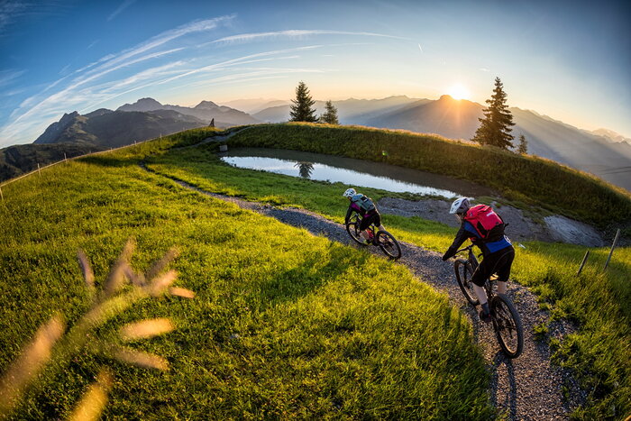 Mountainbiken in Grossarltal | © mtb_travel Two mountain bikers ride a narrow trail through green meadows at sunset with scenic views of the Grossarl Valley. | © mtb_travel