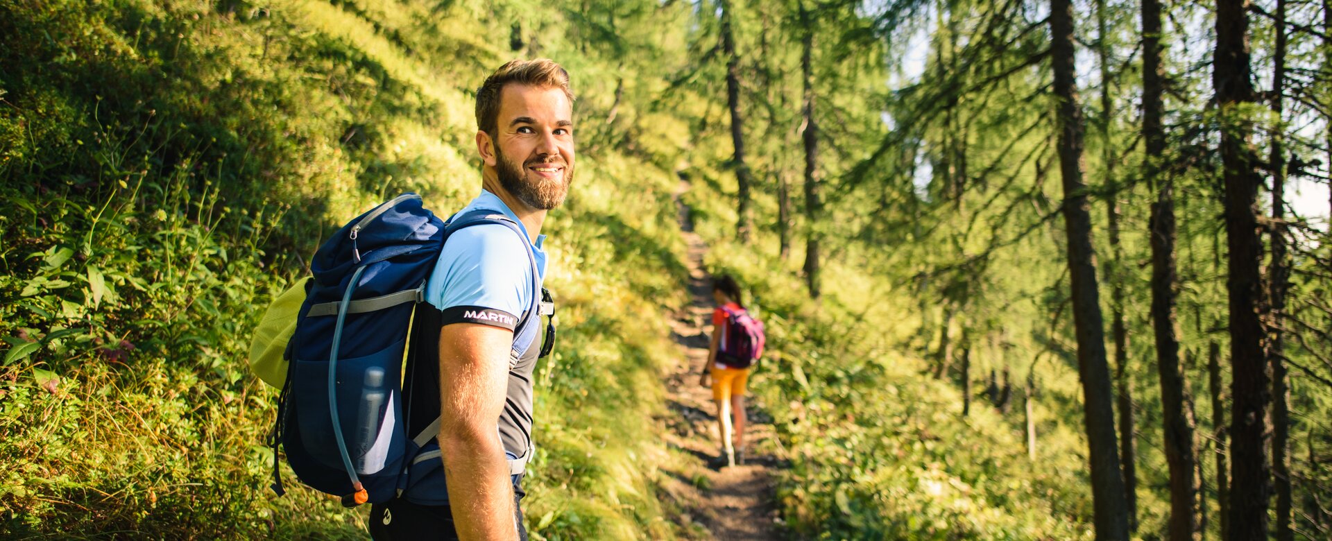 Ein Mann mit Rucksack wandert bei Sonnenschein durch einen lichten Bergwald, schaut lächelnd zurück, im Hintergrund eine Frau | © Salzburger Sportwelt - Christian Schartner