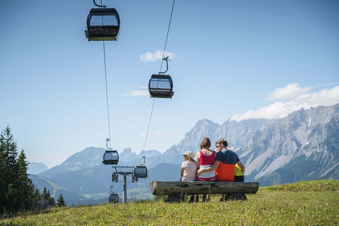 Family enjoys mountain panorama on bench beneath gondolas in Ski amadé A family sits on a wooden bench under gondolas, admiring the impressive mountain panorama in the Austrian region of Ski amadé.