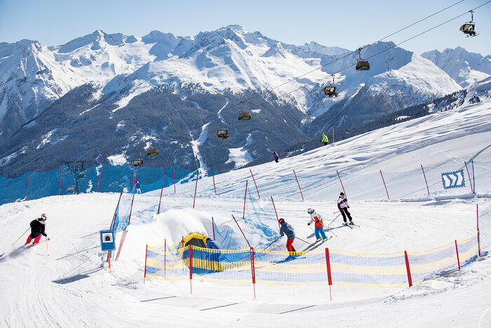 Skiing in Gastein | © Roland Haschka Four people ski on a funslope towards a tunnel and in the background you can see a slope with skiers and above them a chairlift is running | © Roland Haschka